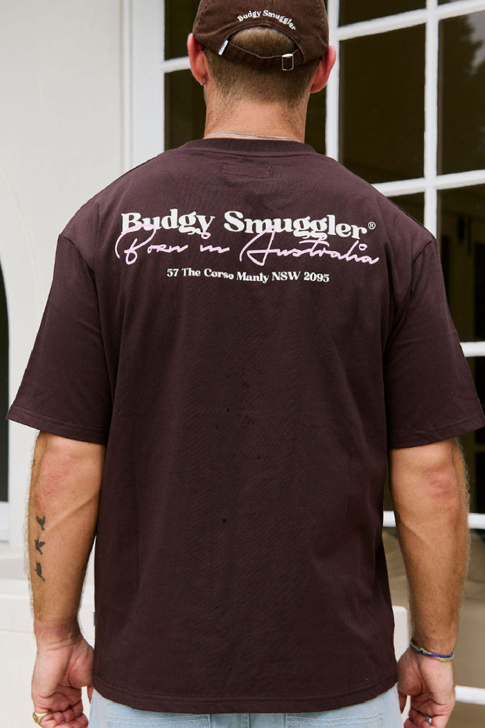 Man wearing a brown t-shirt with 'Budgy Smuggler' branding, standing in front of a building.