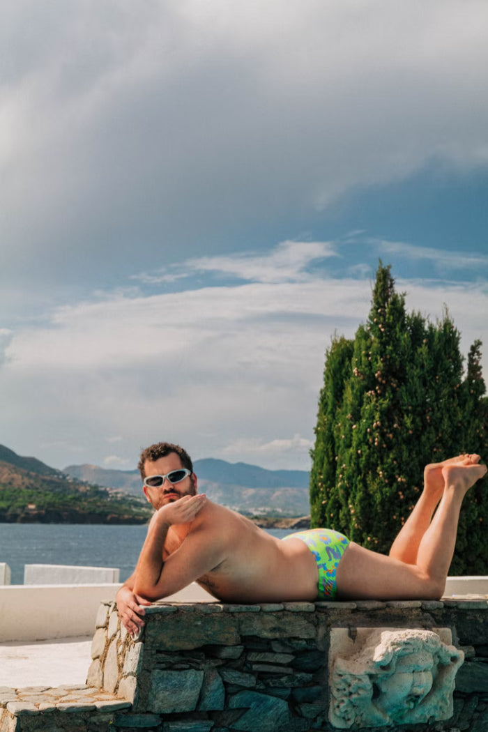 Man lying down wearing green swimwear with a crocodile floral print by the water